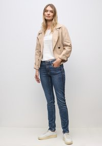 Beżowa marynarka podwójnie zapinana, biała koszula, niebieskie dżinsy z bocznymi paskami i białe sneakersy. Gładka faktura, dopasowany krój.