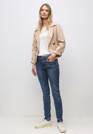 Giacca beige doppiopetto, camicia bianca, jeans blu con strisce laterali e sneakers bianche. Tessuto liscio, design aderente.