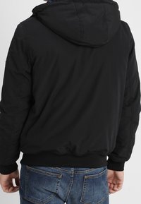 Muž v černé bundě s kapucí a modrých džínách, zobrazený zezadu na jednobílém pozadí.