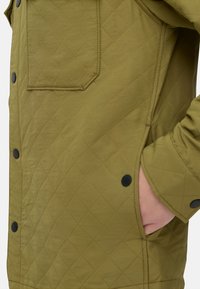 Gequilted olijfgroene jas met een zijzak, zwarte drukknoopsluiting en gestructureerde stof, die een ruitpatroon en een gladde afwerking toont.