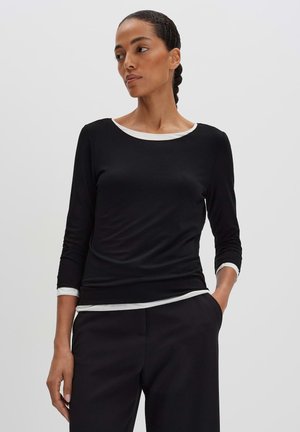 Donna con capelli intrecciati indossa una maglia nera a maniche lunghe sopra una canottiera bianca e pantaloni neri, in piedi con una mano in tasca, guarda di lato.