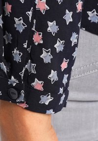 Camicia button-up in tessuto navy con motivo a stelle rosa, bianche e blu. Dettaglio delle maniche a polsino con un bottone nero visibile.