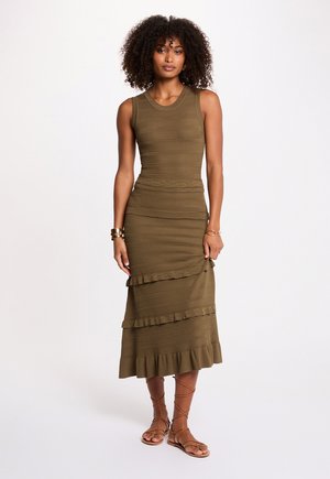 Kvinde med krøllet hår iført ærmeløs olivengrøn ribstrikket kjole og brune flade sandaler med remme, stående mod hvid baggrund.