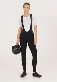 Zwarte fiets-bibshorts met mesh schouderbanden, gecombineerd met een wit textuurshirt. Een zwarte helm wordt in de linkerhand gehouden.