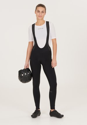 Zwarte fiets-bibshorts met mesh schouderbanden, gecombineerd met een wit textuurshirt. Een zwarte helm wordt in de linkerhand gehouden.