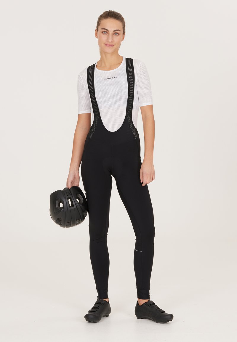 Zwarte fiets-bibshorts met mesh schouderbanden, gecombineerd met een wit textuurshirt. Een zwarte helm wordt in de linkerhand gehouden.