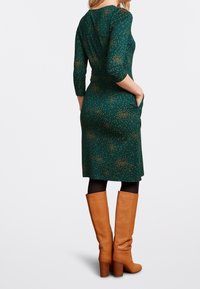 Grön klänning med trekvartsärmar, prydd med ett mönster av guldprickar, tillsammans med knähöga bruna läderstövlar och svarta strumpbyxor.