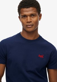 Hombre con cabello corto y rizado que lleva una camiseta azul marino de SuperDry con un logo rojo en el pecho, mirando hacia adelante contra un fondo blanco liso.