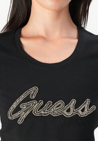 Musternäriga must T-särk ümmarguse kaelusega, millel on rhinestone'i kaunistatud "Guess" logo ja kontrastse värviga "est.1981" detail.