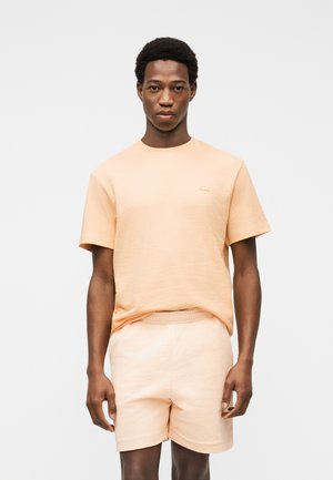 Joven de pie de frente vistiendo una camisa de manga corta y pantalones cortos a juego de color durazno sobre un fondo blanco liso.