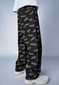 Černé džíny z denimu s bílým nápisem ve stylu graffiti. Volný střih s zadní kapsou, nošené s bílými teniskami.