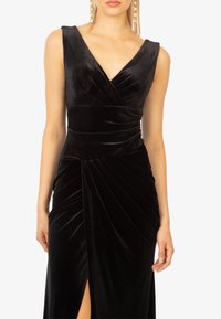 Abito in velluto nero con scollo a V profondo, plissettatura in vita e spacco alta coscia, che mette in mostra una texture liscia e ricca.