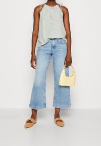 Lichtgroene, patroonrijke mouwloze top met strikbandjes, blauwe wijde jeans, beige platte schoenen en een kleine gele handtas.