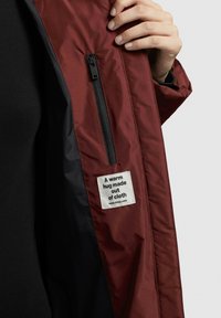 Chaqueta burdeos con un bolsillo con cremallera negra y una etiqueta que dice "Un abrazo cálido hecho de tela." Textura de tela suave y lisa visible.