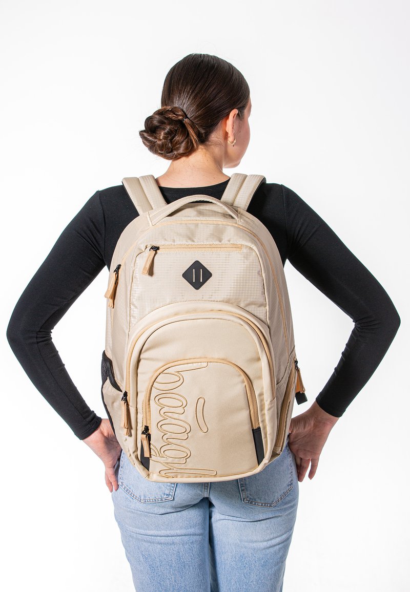 Zaino beige con tessuto testurizzato, tasche con doppia zip e un design in rilievo frontale. Presenta spallacci regolabili e una patch a forma di diamante.