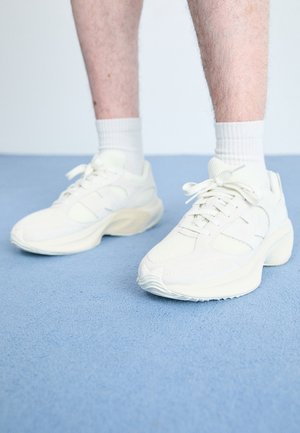 Person, die weiße klobige Sneaker und weiße Knöchelsocken trägt, steht auf einem hellblauen strukturierten Boden.