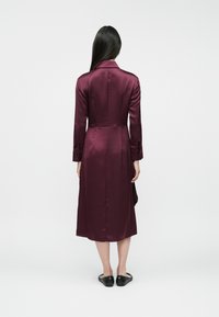 Bordowy satynowy długi płaszcz z ogonem, z długimi rękawami, kołnierzem i zapinanymi mankietami, noszony na czarnych butach wsuwanych.