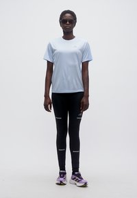 Γυναίκα που φοράει ανοιχτό μπλε αθλητικό μπλουζάκι, μαύρο κολάν με ανακλαστικές ρίγες, μοβ παπούτσια για τρέξιμο και σκούρα γυαλιά ηλίου, στέκεται μπροστά σε λευκό φόντο.