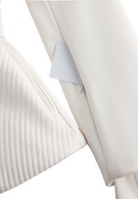 Jachetă albă cu textură matlasată pe mâneca stângă, design elegant și o mică etichetă albă parțial vizibilă lângă fermoar.