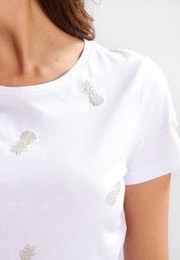 Bílý bavlněný tričko s malými vyšívanými stříbrnými ananasy. Kulatý výstřih a krátké rukávy s hladkým povrchem.