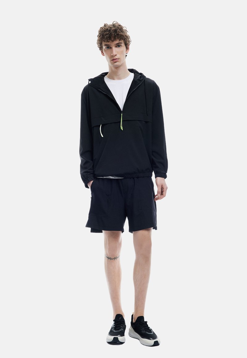 Chaqueta ligera negra con capucha y cordones verdes, combinada con pantalones cortos oscuros y zapatillas deportivas negras, con un ajuste casual.
