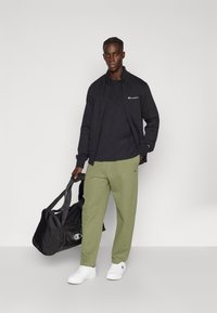 Μαύρο μπουφάν με φερμουάρ πάνω από μαύρη μπλούζα, πράσινες φόρμες και λευκά αθλητικά παπούτσια. Κρατάει μαύρη τσάντα ταξιδιού με λουριά ώμου.