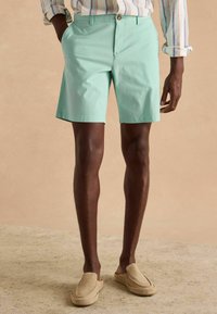 Mint zaļas šortus ar plakanu priekšpusi, aizmugures kabatām un pogu aizdari, apvienojot ar smilšu krāsas apaviem bez šņorēm. Rādīta teksturēta auduma virsma.