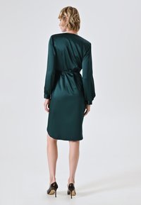 Satin-Kleid in smaragdgrün mit langen Ärmeln, tailliert und mit subtiler glänzender Oberfläche; knielang mit gewelltem Saum. Dazu schwarze Stiletto-Highheels.