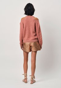Roze oversized trui met cold shoulder ontwerp, gecombineerd met metallic bronzen shorts. Textuurgebreide en glanzende stofdetails.