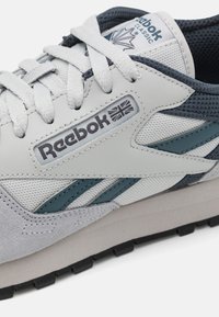 Klasické tenisky Reebok v šedé a námořnické modři, s texturovaným svrškem z kůže a síťoviny, plochými tkaničkami a světle šedou podrážkou s černým vzorkem.
