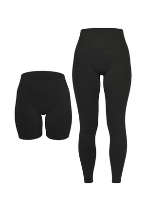 Frau trägt schwarze Leggings mit hohem Bund, ein passendes schwarzes Crop-Top und weiße Sportschuhe, steht mit den Händen an den Seiten.