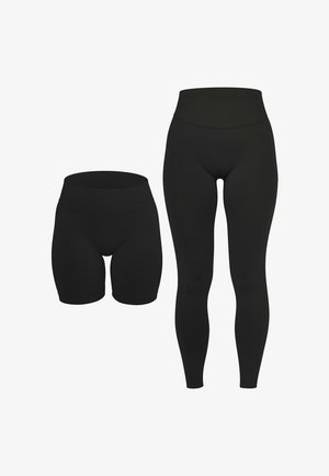 Frau trägt schwarze Leggings mit hohem Bund, ein passendes schwarzes Crop-Top und weiße Sportschuhe, steht mit den Händen an den Seiten.