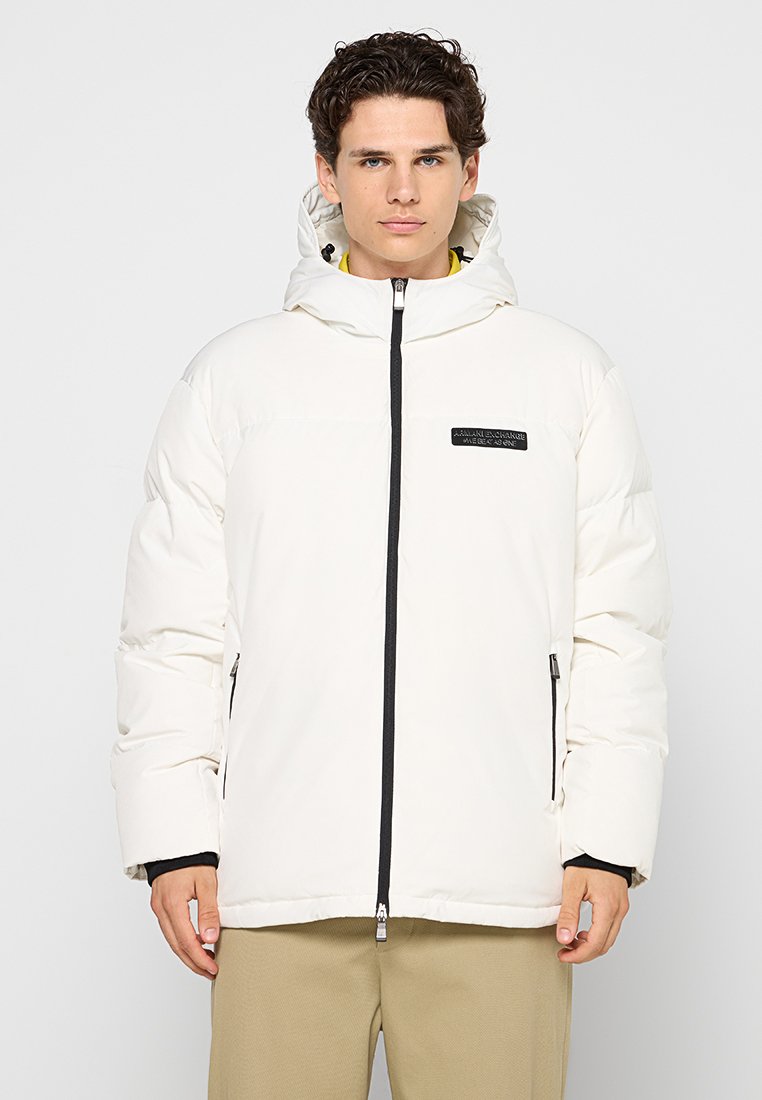 Giacca puffer bianca con chiusura a zip nera, cappuccio e patch con logo sul petto. Presenta tasche laterali e polsini elasticizzati.