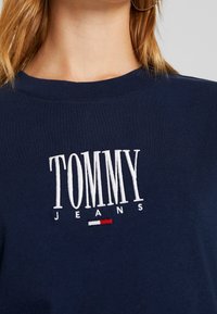 Marinblå bomullströja med rund halsringning, prydd med vit broderad text "TOMMY" och "JEANS", samt en liten röd och vit detalj nedanför.