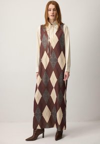 Obleka brez rokavov v vzorcu diamantov v barvah bordo rdeče, kreme in sive, z visokim ovratnikom in sproščenim krojem, kombinirana s rjavimi škornji s peto.