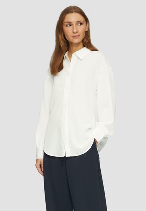 Chemise blanche à manches longues avec une finition texturée, dotée d'un col et d'une coupe décontractée. Associée à un pantalon sombre.