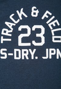 Tela azul marino con texto blanco "PISTA Y CAMPO" en arco sobre el número "23" y texto parcialmente visible "S-DRY. JPN" debajo.