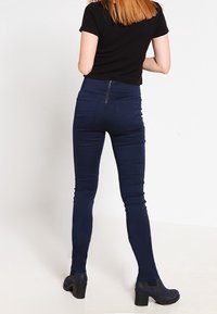 Námořnické modré skinny džíny s vysokým pasem, hladkou strukturou a zadním zipem. Nosené s černým topem s krátkým rukávem a černými vysokými botami.