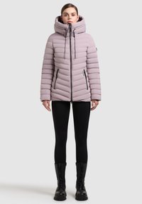 Lys lilla oppustelig jakke med hætte, lynlås lukning og sidelommer. Har et chevronmønster og justerbare snore. Sammen med sorte leggings og støvler.