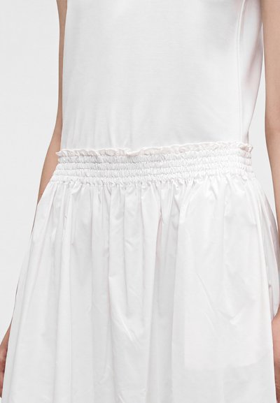 Gros plan d'une personne portant un haut blanc sans manches rentré dans une jupe blanche avec une ceinture élastiquée froncée.
