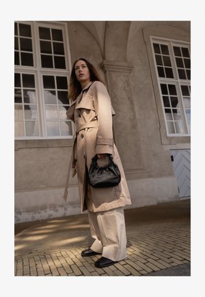 Femme en trench-coat beige et pantalon large tenant un sac à main noir, debout dans un couloir voûté avec de grandes fenêtres et un sol en briques à motif.