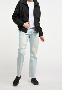 Personne portant une veste noire zippée, un t-shirt blanc, un jean bleu clair déchiré et des baskets noires, debout devant un fond uni.