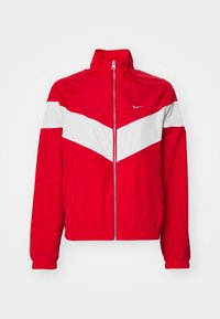 Chaqueta de chándal roja de Nike con una franja en chevron blanca en el pecho, cierre de cremallera en la parte delantera, puños elásticos y cuello alto.