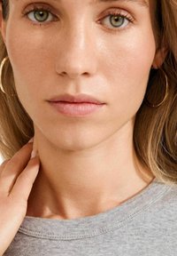 Orecchini a cerchio d'oro, scollo di una felpa grigia, pelle liscia ed espressione neutra. I capelli cadono morbidi sulla spalla.