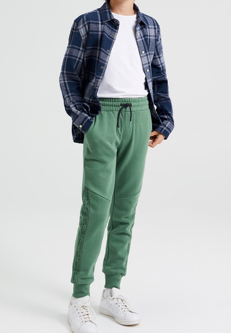 Gröna sweatpants med dragsko i midjan, strukturerad tyg och sidopaneler. Matcher med en blå rutig skjorta över en vit t-shirt.