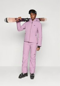 Slēpošanas tērps gaiši violetā krāsā, kas sastāv no rāvējslēdzēja jaciņas un atbilstošiem biksēm. Kalnu slēpju apavi un slēpes tiek turēti pār plecu. Virsma šķiet gluda.