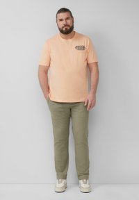 Helles Pfirsich-Baumwoll-T-Shirt mit Frontgrafik und kurzen Ärmeln, kombiniert mit olivgrünen Hosen und weißen Turnschuhen. Lässiger Schnitt.