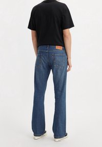 Pantalones vaqueros de mezclilla azul de campana con un acabado ligeramente desgastado, que cuentan con un parche de cuero en la parte trasera, bolsillos estándar y zapatillas negras.