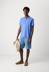 Modrá polokošeľa s bielym lemom, džínové šortky a hnedé dreváky. Modelka drží svetlo béžový kabát. Jednoduchý, neformálny outfit.