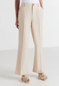 Pantaloni beige a vita alta e gamba larga con pieghe frontali, tasche laterali, chiusura con bottone e zip, indossati con tacchi aperti beige decorati con fiori.